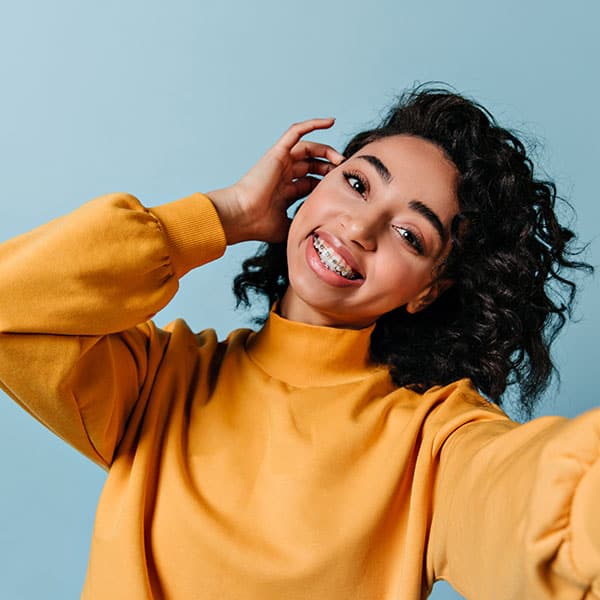 Woman with braces smiling at camera, wearing a yellow sweatshirt against a light blue backdrop, taking a selfie-like photo.