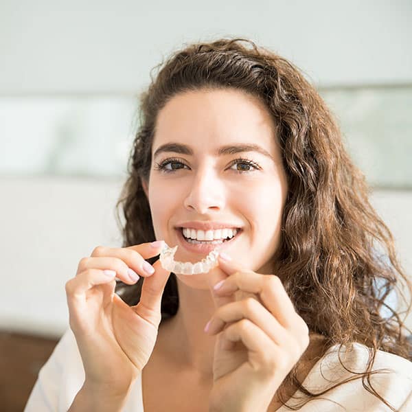 Woman smiling while holding a clear dental aligner up to her teeth, showing it off between her fingers.
