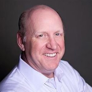 Smiling man in a light blue shirt posing for a headshot against a dark gray background, showing shoulders and upper body.
